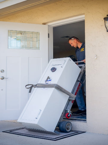 A large white safe, strapped to a dolly and being unloaded from a house by two movers. One mover stands outside on the porch while the other pushes the dolly up the steps into the home.